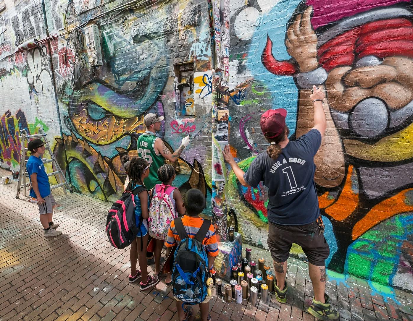 A group of children watching two graffiti artists spray paint a mural on graffiti alley